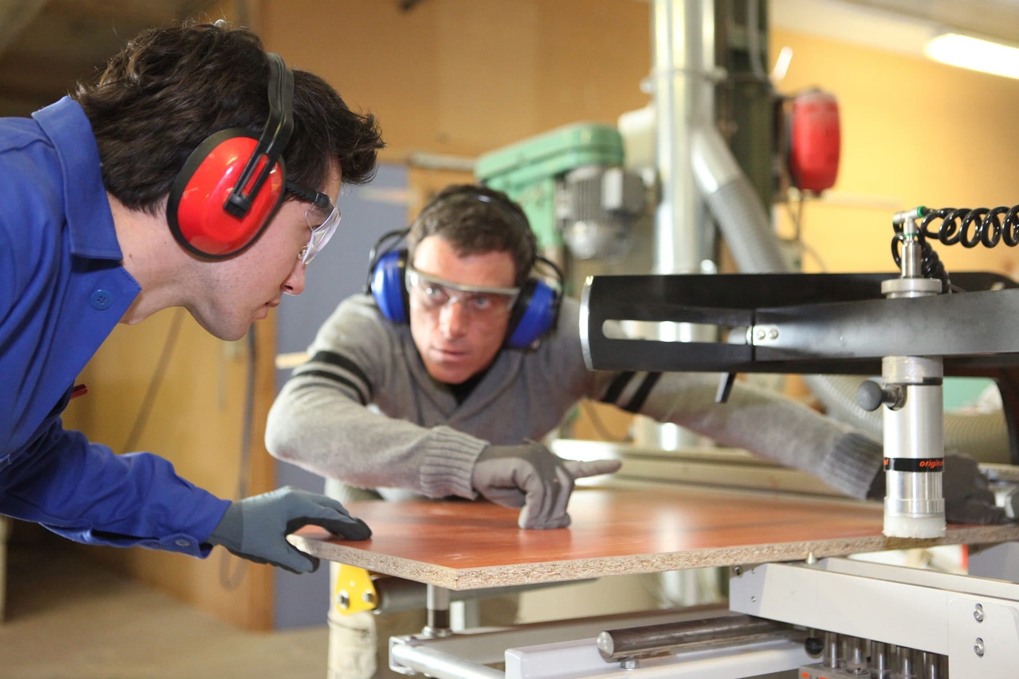 An Experienced Worker Showing An Apprentice How To Cut A Piece Of Wood Using A Machine