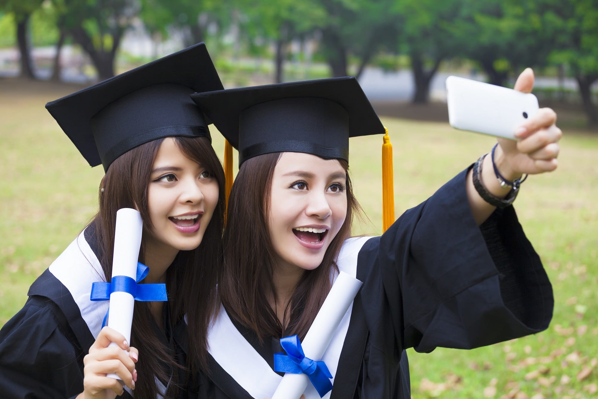 Two Happy Women In Graduation Gowns Taking Picture With Cell Pho