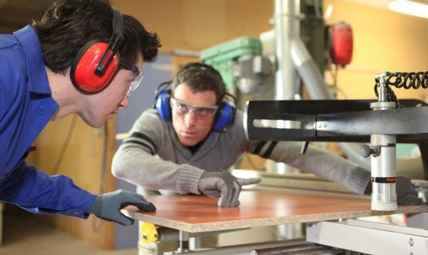 An Experienced Worker Showing An Apprentice How To Cut A Piece Of Wood Using A Machine