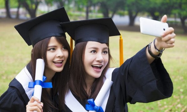 Two Happy Women In Graduation Gowns Taking Picture With Cell Pho